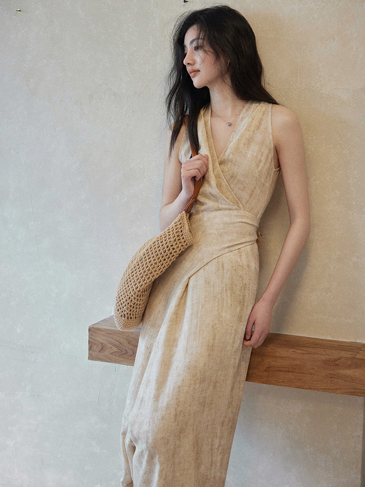 A woman in a beige wrap dress with a V-neck and asymmetrical hemline, holding a woven bag.