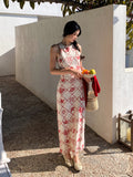 A woman in a long, sleeveless dress with a red diamond grid pattern and floral motifs stands outdoors holding a bowl of fruit.