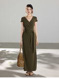 A woman in an olive green wrap dress with a V-neck and tie belt, holding a woven bag.