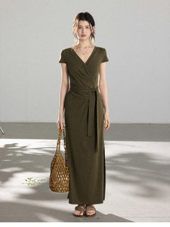 A woman in an olive green wrap dress with a V-neck and tie belt, holding a woven bag.