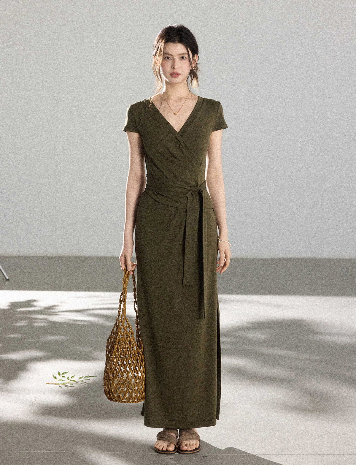 A woman in an olive green wrap dress with a V-neck and tie belt, holding a woven bag.