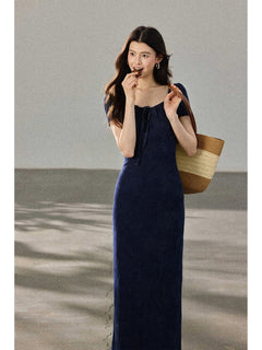 A woman in a navy blue fitted long dress with a square neckline and drawstring detail, holding a woven tote bag.