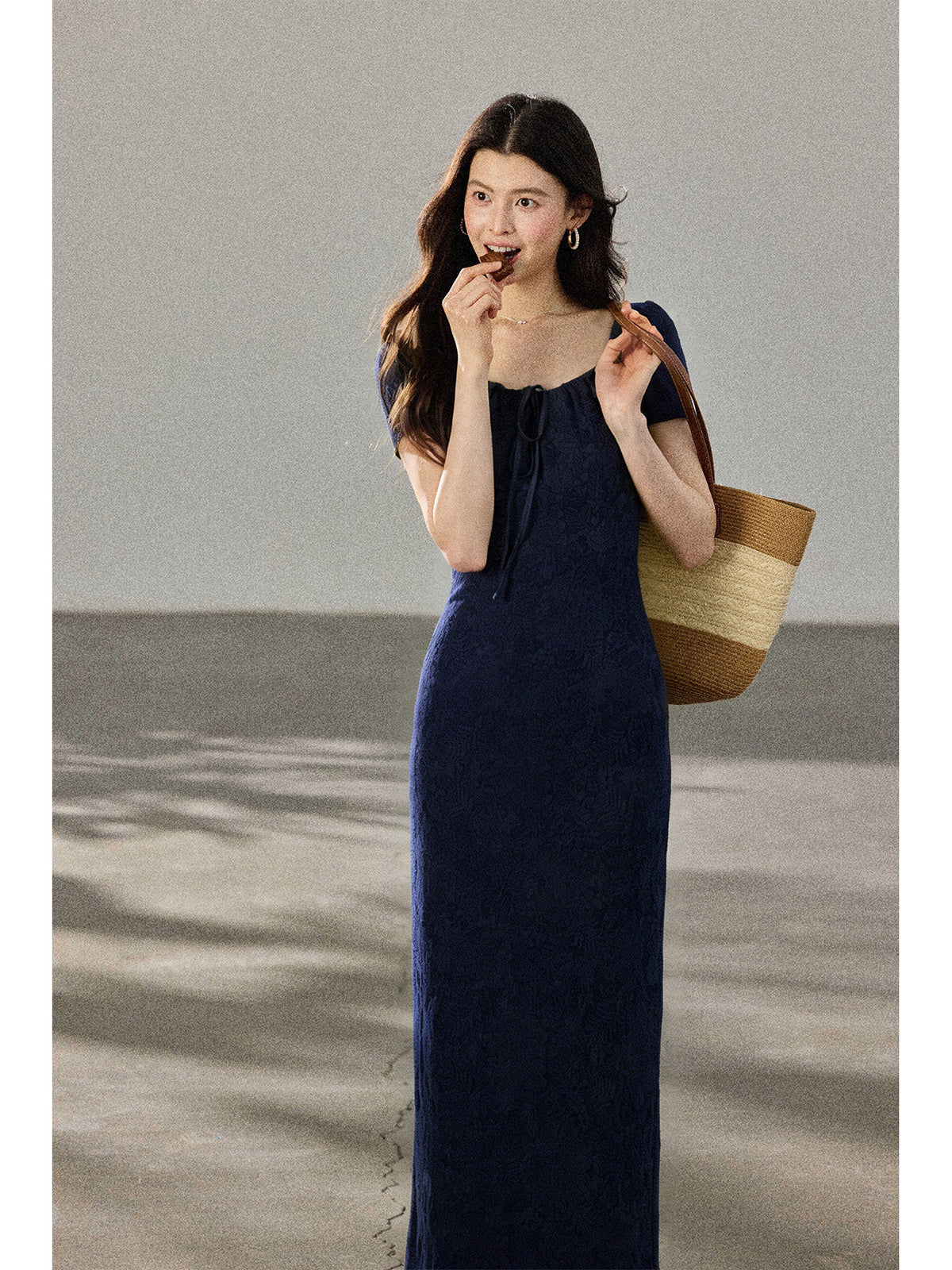 A woman in a navy blue fitted long dress with a square neckline and drawstring detail, holding a woven tote bag.