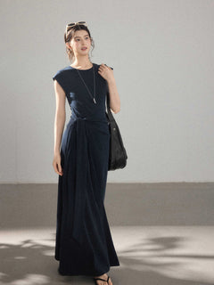 A woman wearing a navy blue long A-line dress with a waist tie and side pleats.