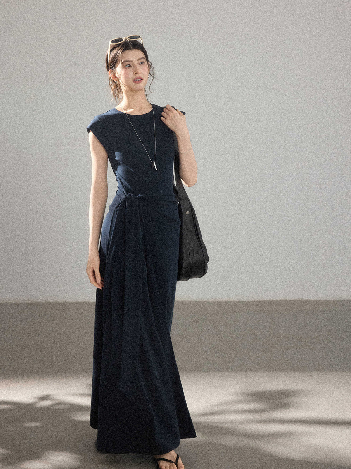 A woman wearing a navy blue long A-line dress with a waist tie and side pleats.