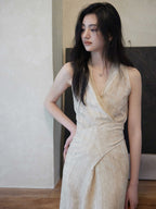 A woman in a beige wrap dress with a V-neck and asymmetrical hemline, standing against a textured wall.