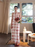 A woman in a long, sleeveless dress with a red diamond grid pattern and floral motifs stands indoors holding a bouquet of roses.