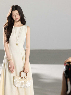 A woman in a cream sleeveless A-line floor-length dress, holding a round sequined handbag.