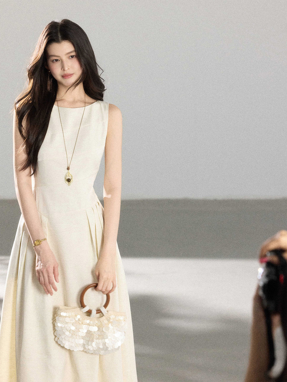 A woman in a cream sleeveless A-line floor-length dress, holding a round sequined handbag.