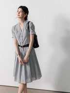 A woman in a grey A-line midi dress with a V-neck collar and pleated skirt, standing against a white wall.