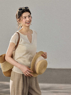 A woman holds a straw hat and bag while wearing a cream-colored knitted top with a V-neck and ribbed trim at the hem.