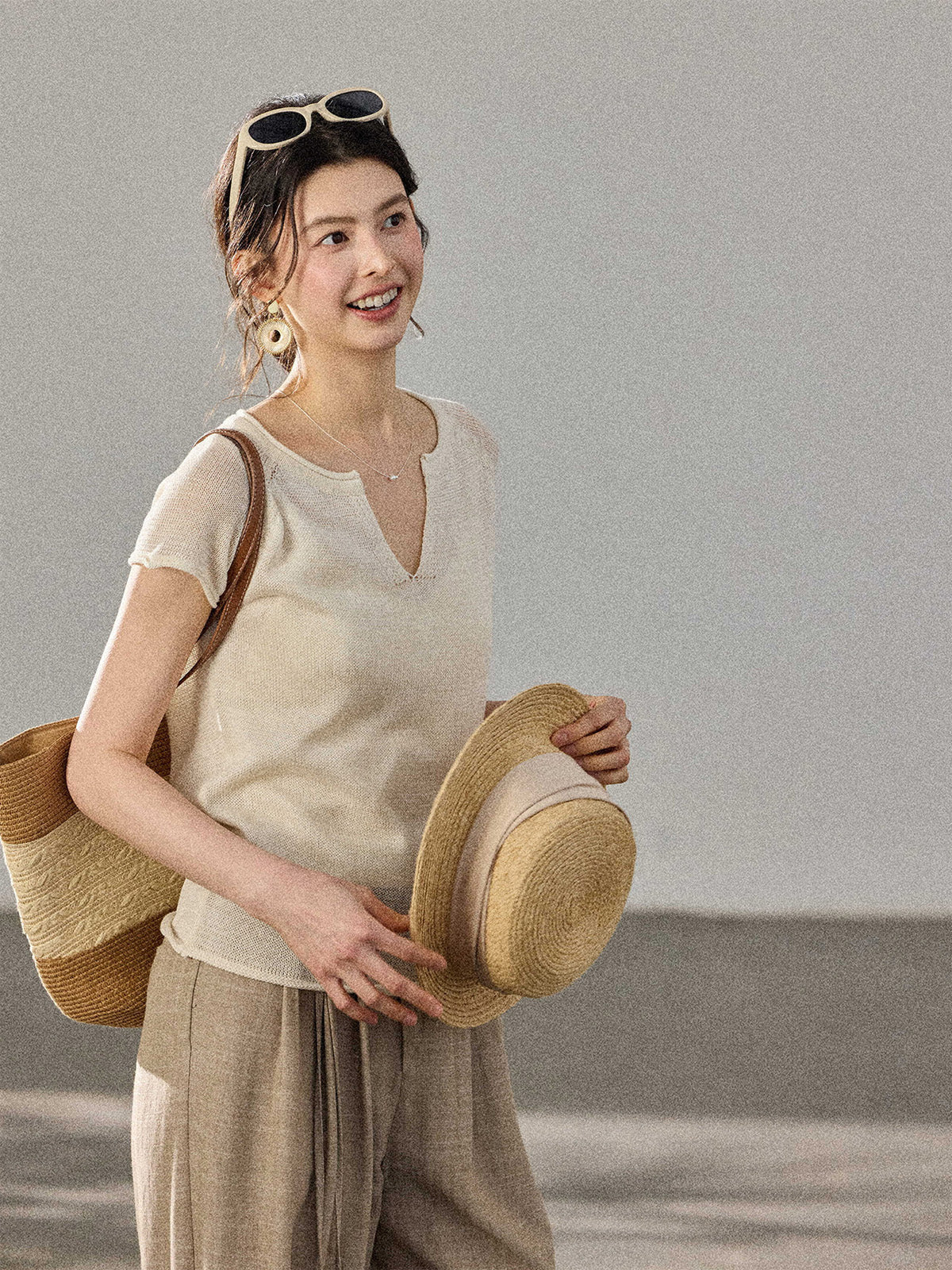 A woman holds a straw hat and bag while wearing a cream-colored knitted top with a V-neck and ribbed trim at the hem.