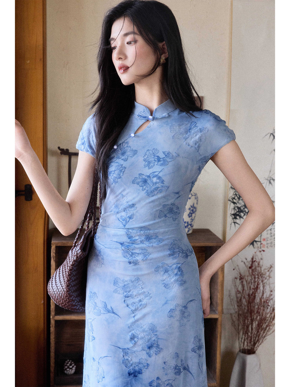 A woman in a light blue floral dress with a mandarin collar and keyhole opening, standing while holding a woven bag.