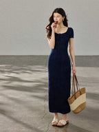A woman in a navy blue long dress with a square neckline, standing on a concrete surface while holding a woven tote bag.