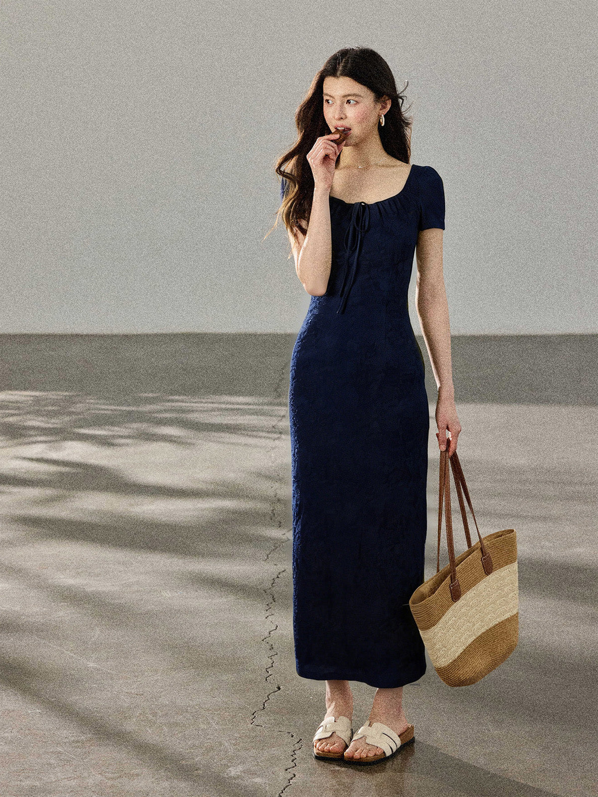 A woman in a navy blue long dress with a square neckline, standing on a concrete surface while holding a woven tote bag.