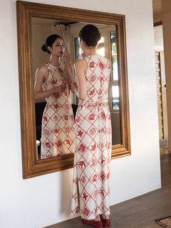 A woman in a long, sleeveless dress with a red diamond grid pattern and floral motifs stands before a mirror.