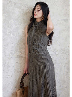 A woman in a sleeveless black gingham floor-length dress with a high neck and tie detail, holding a woven bag.