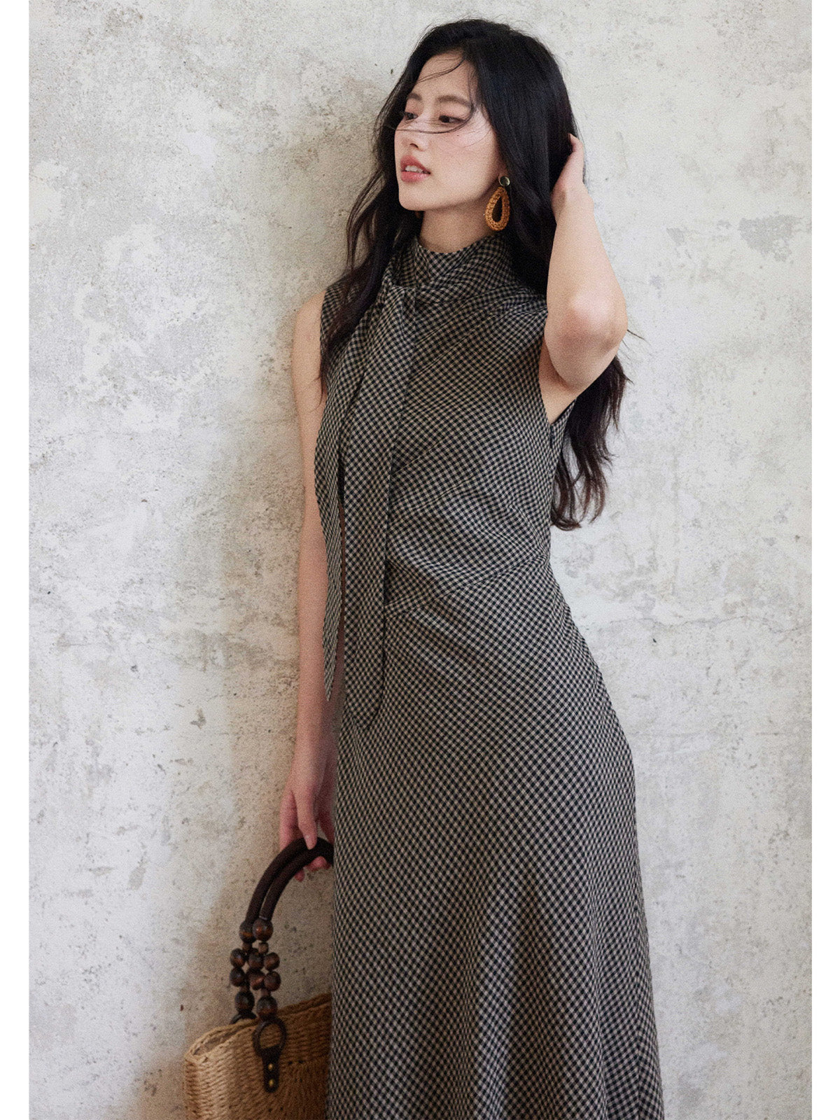 A woman in a sleeveless black gingham floor-length dress with a high neck and tie detail, holding a woven bag.