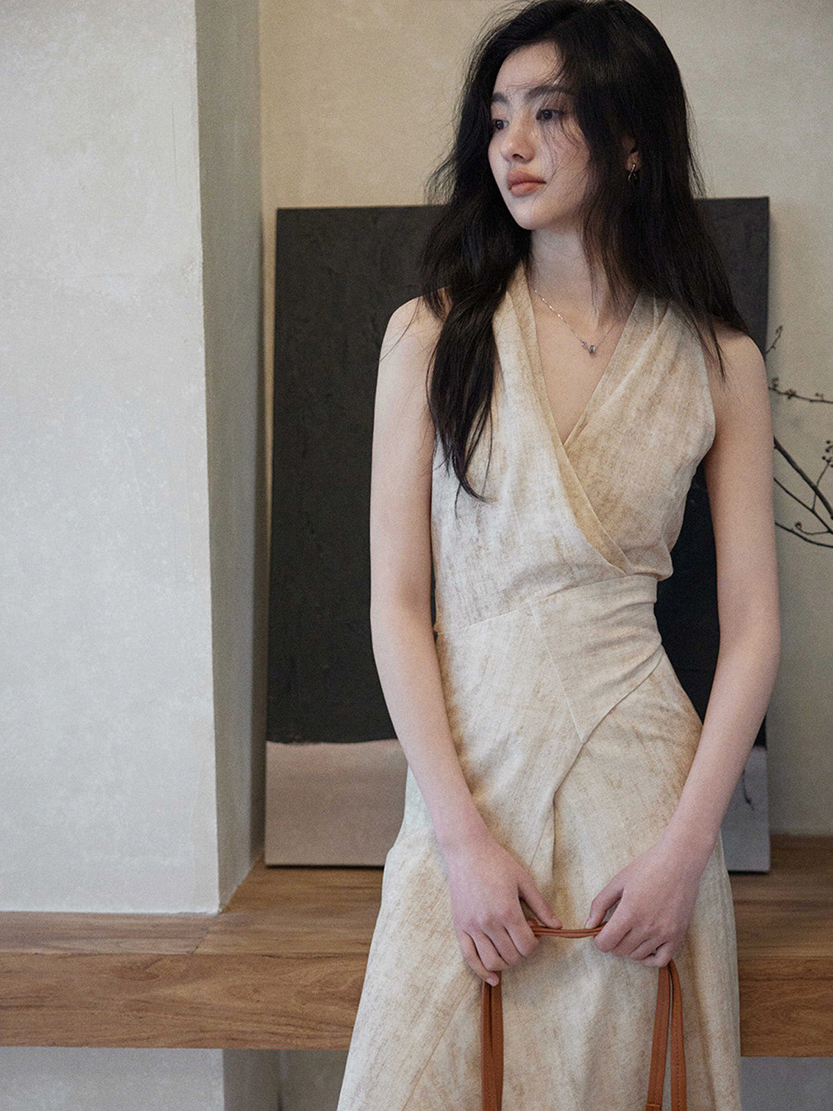 A woman in a beige wrap dress with a V-neck and asymmetrical hemline, standing while holding a woven bag.