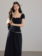 A woman wearing a black long dress with a square neckline, puff sleeves, and a metallic chain belt.