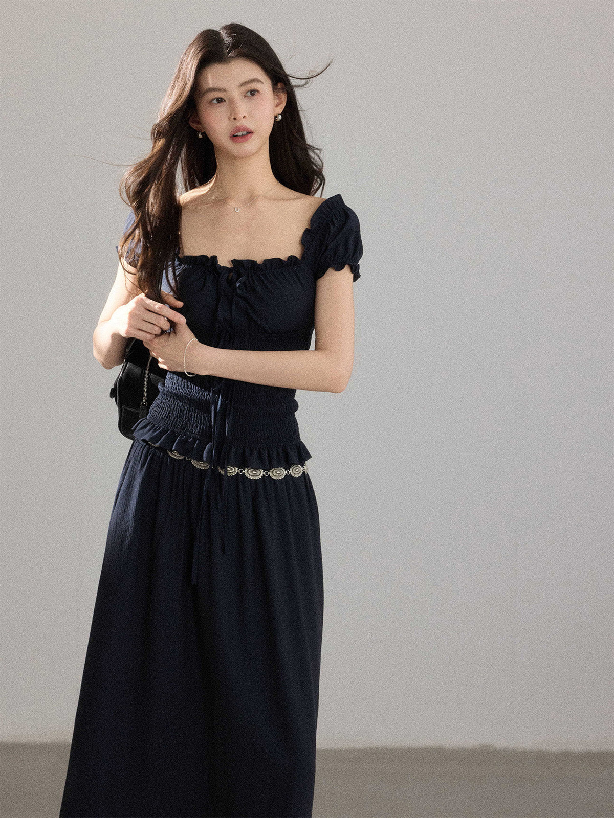 A woman wearing a black long dress with a square neckline, puff sleeves, and a metallic chain belt.