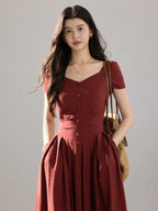 A woman in a red dress with a V-neck and pleated skirt, standing against a grey background.