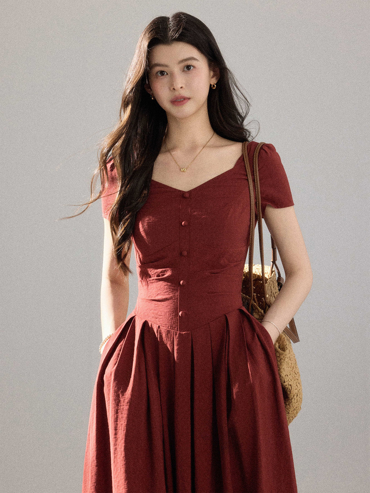 A woman in a red dress with a V-neck and pleated skirt, standing against a grey background.