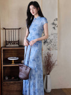 A woman in a light blue floral dress with a mandarin collar and keyhole opening, standing next to a wooden shelf.