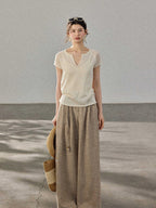 A woman stands in profile wearing a cream-colored knitted top with a V-neck and ribbed trim at the hem.