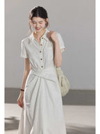 A woman wearing a white A-line dress with a button-front, collar, and twisted waist tie.