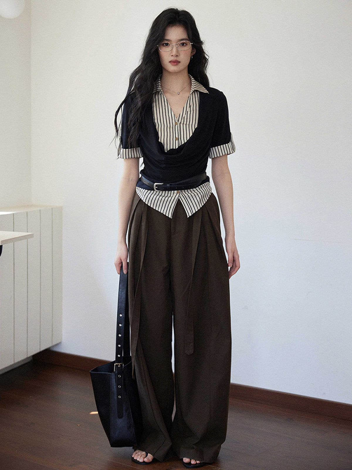 A woman stands in a black and white striped layered V-neck top with short sleeves, paired with brown pants.