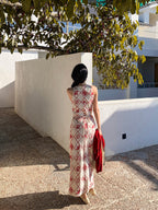A woman in a long, sleeveless dress with a red diamond grid pattern and floral motifs walks away from the camera.