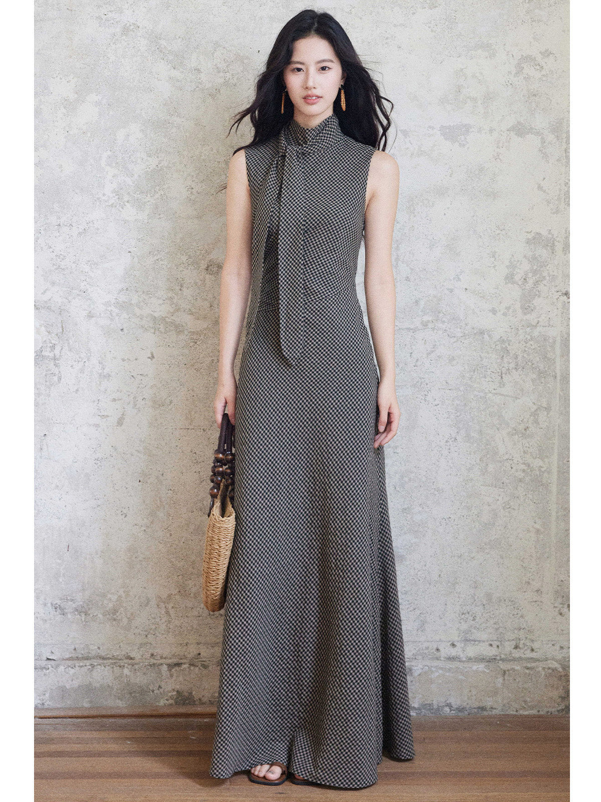 A woman in a sleeveless black gingham floor-length dress with a high neck and tie detail, holding a woven bag.