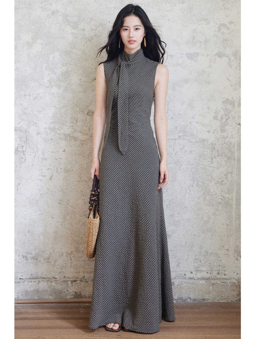 A woman in a sleeveless black gingham floor-length dress with a high neck and tie detail, holding a woven bag.