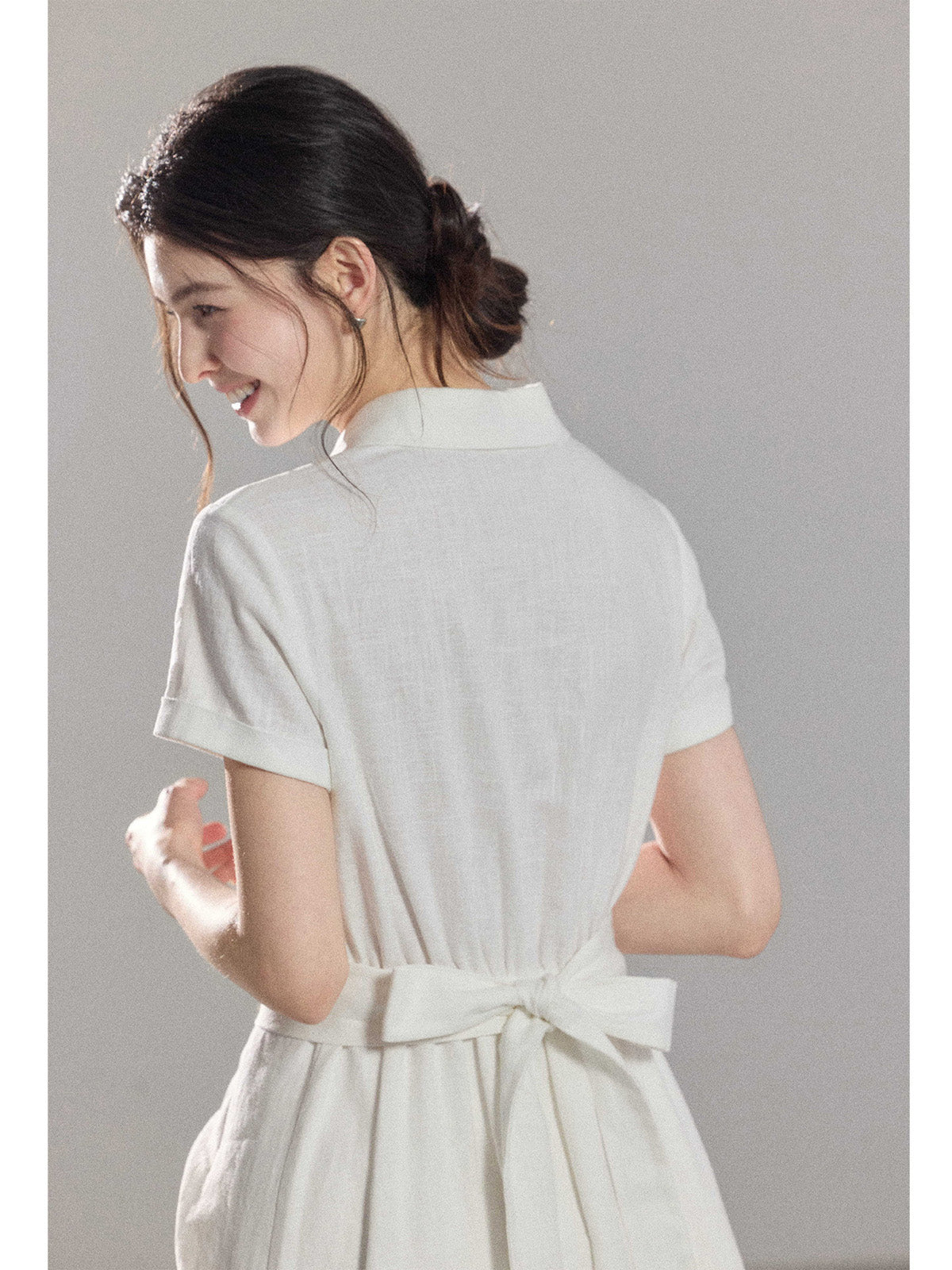 A woman wearing a white A-line dress with a button-front, collar, and twisted waist tie.