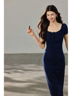 A woman in a navy blue fitted long dress with a square neckline and drawstring detail, walking while holding a chocolate bar.