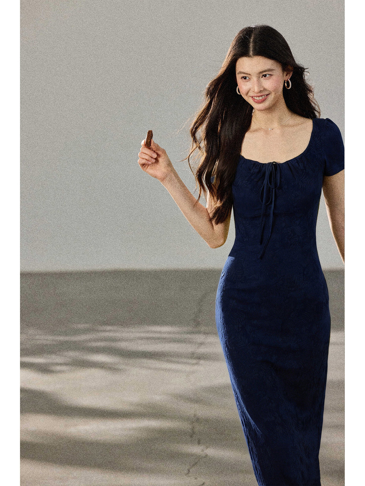 A woman in a navy blue fitted long dress with a square neckline and drawstring detail, walking while holding a chocolate bar.