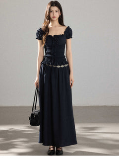 A woman wearing a black long dress with a square neckline, puff sleeves, and a metallic chain belt.