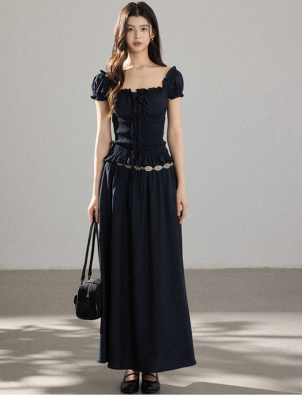 A woman wearing a black long dress with a square neckline, puff sleeves, and a metallic chain belt.