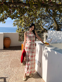 A woman in a long, sleeveless dress with a red diamond grid pattern and floral motifs stands on a terrace overlooking the sea.