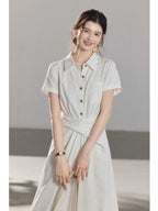 A woman wearing a white A-line dress with a button-front, collar, and twisted waist tie.