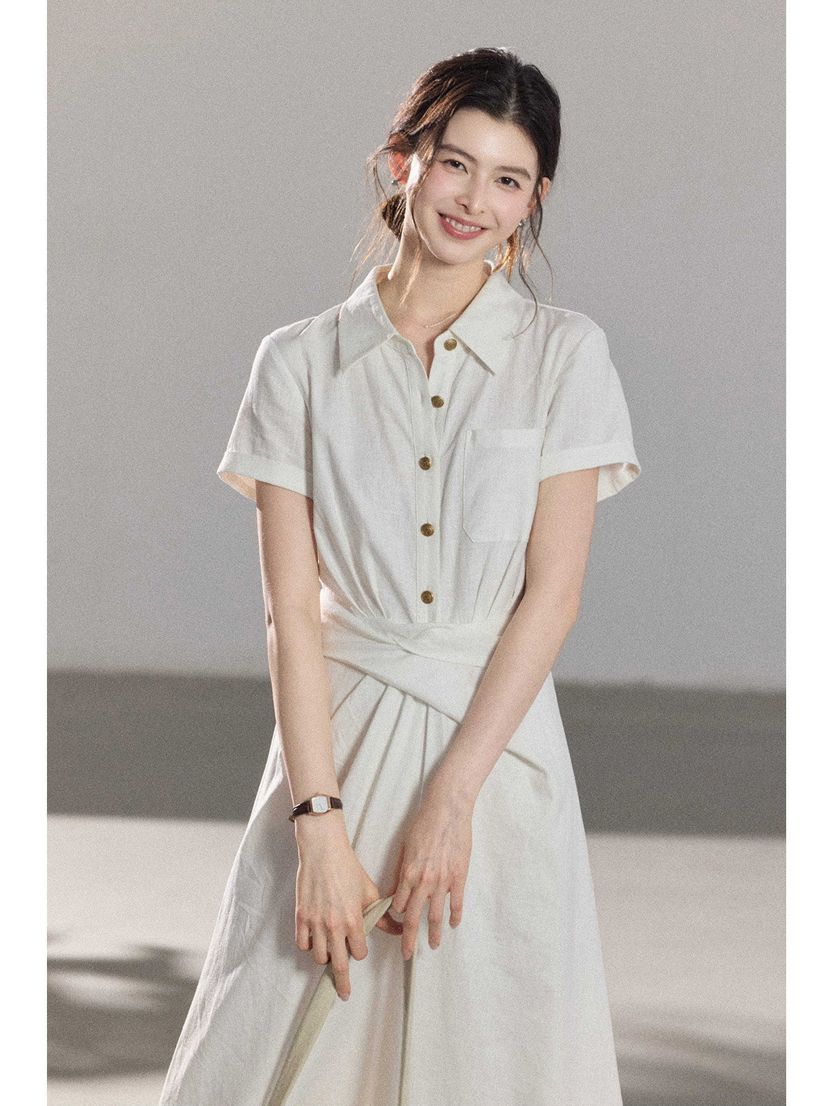 A woman wearing a white A-line dress with a button-front, collar, and twisted waist tie.
