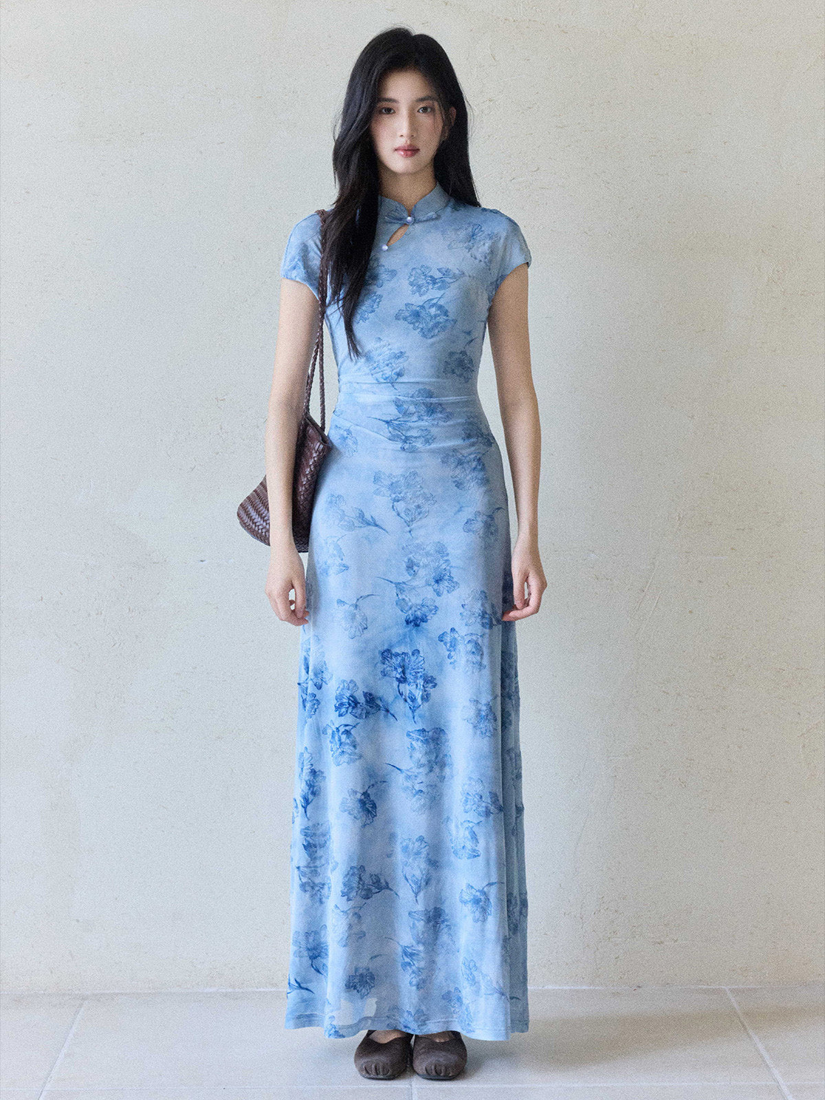 A woman in a light blue floral dress with a mandarin collar and keyhole opening, standing against a plain wall.