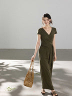 A woman in an olive green wrap dress with a V-neck and tie belt, smiling while holding a woven bag.