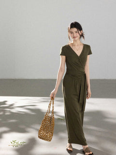 A woman in an olive green wrap dress with a V-neck and tie belt, smiling while holding a woven bag.