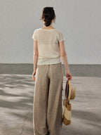 A woman stands with her back to the camera, wearing a cream-colored knitted top with a V-neck and ribbed trim at the hem.