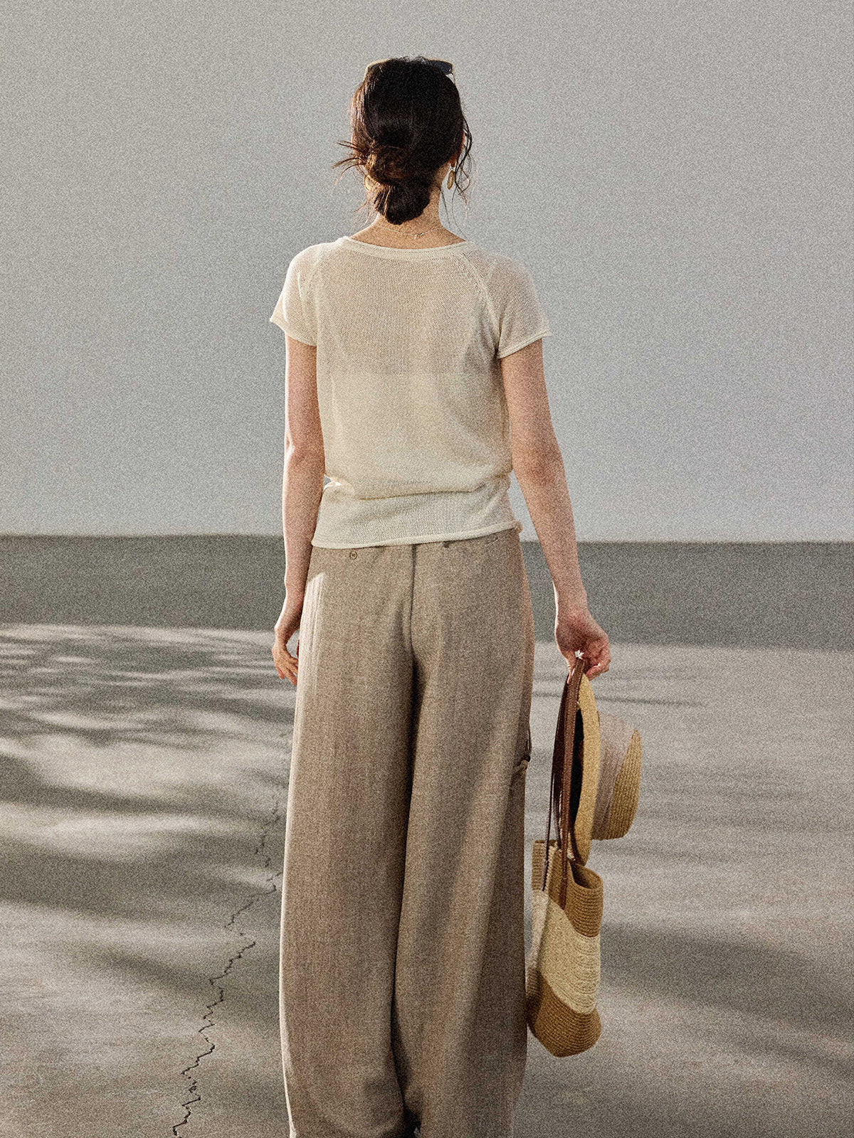 A woman stands with her back to the camera, wearing a cream-colored knitted top with a V-neck and ribbed trim at the hem.