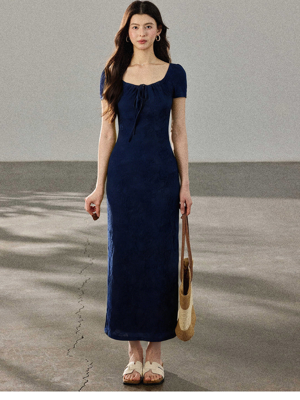 A woman in a navy blue fitted long dress with a square neckline and drawstring detail, standing on a concrete surface.