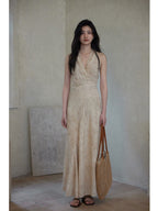A full-length view of a woman in a beige wrap dress with a V-neck and asymmetrical hemline, holding a woven bag.