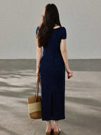 A woman in a navy blue fitted long dress with a square neckline and drawstring detail, viewed from the back showing a side slit.